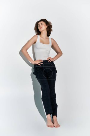 Photo for A pretty young woman poses gracefully in a white tank top and black pants against a grey studio backdrop. - Royalty Free Image