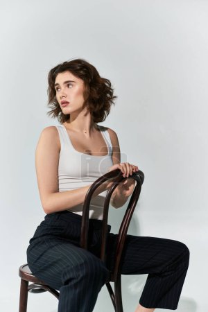 A pretty young woman poses gracefully on a wooden chair, exuding elegance and confidence in a studio setting.