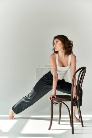 A pretty young woman in black pants and white tank top sits gracefully atop a wooden chair in a studio setting against a grey background.
