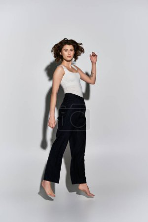 A pretty young woman posing gracefully in a white tank top and black pants in a studio setting against a grey background.