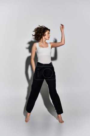 Foto de Una mujer joven y bonita toma una pose elegante con pantalones negros y camiseta blanca contra un fondo gris del estudio. - Imagen libre de derechos