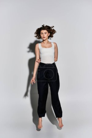 A pretty young woman in black pants and white tank top poses gracefully in front of a stark white wall in a studio setting.
