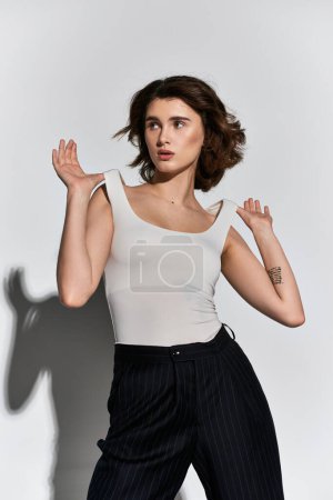 A young woman stands gracefully in front of a blank white wall, exuding elegance and confidence in her black pants and white tank top.