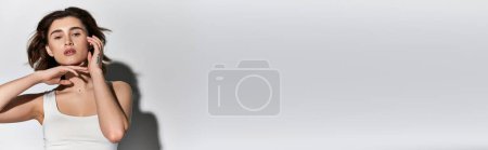 Téléchargez les photos : Une jeune femme élégante dans un débardeur blanc tient une main à son oreille dans un studio sur un fond gris. - en image libre de droit