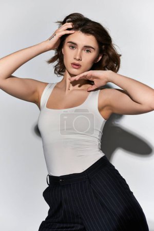 A pretty young woman in black pants and a white tank top strikes a stylish pose in a studio against a grey background.
