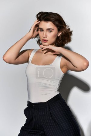 A pretty young woman posing confidently in a white tank top and black pants in a studio setting on a grey background.