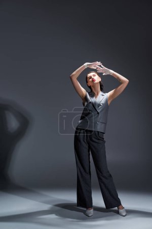 Foto de Mujer joven con estilo en un chaleco gris y pantalones golpea una pose en un entorno de estudio sobre un fondo gris. - Imagen libre de derechos