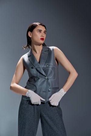 Photo for A stylish young woman strikes a pose in a gray suit with a vest, complemented by white gloves, set against a grey background. - Royalty Free Image