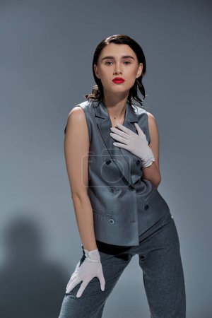 Téléchargez les photos : Jeune femme élégante pose dans un élégant costume gris avec gilet et gants blancs sur un fond de studio gris. - en image libre de droit