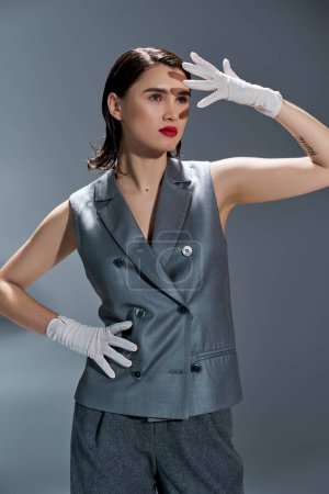 Stylish young woman striking a pose in an elegant gray suit with a vest, complemented by white gloves, on a gray studio background.