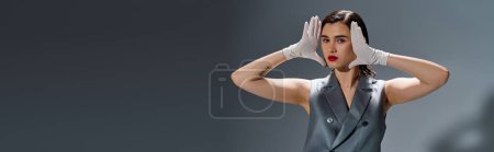 Photo for A stylish young woman poses in an elegant suit with a vest, hands on her head, expressing emotions in a studio setting on a grey background. - Royalty Free Image