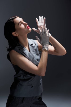 A stylish young woman in an elegant suit with a vest raising her hands in the air in a studio setting against a grey background.