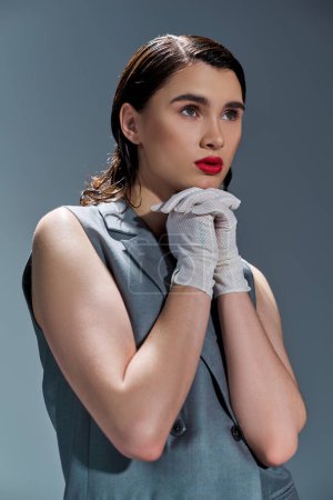 A stylish young woman poses in an elegant gray dress and white gloves in a studio setting on a grey background.
