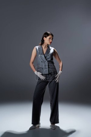 A stylish young woman elegantly poses in a gray suit with a vest and white gloves in a studio setting on a grey background.
