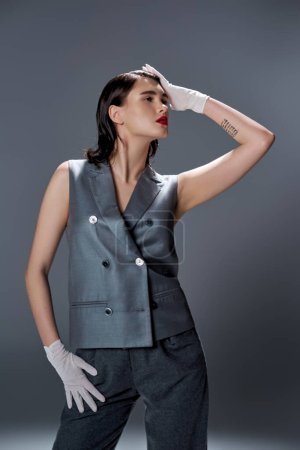 Stilvolle junge Frau posiert in elegantem grauen Anzug mit Weste und weißen Handschuhen, verströmt Anmut und Raffinesse in einem Studio-Setting.