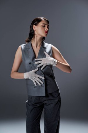 Stilvolle junge Frau posiert im eleganten grauen Anzug mit Weste und weißen Handschuhen in einem Studio vor grauem Hintergrund.