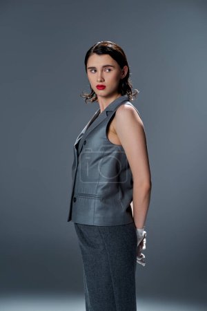 A stylish young woman strikes a pose in an elegant gray suit with a vest, accentuated by bold red lipstick, against a grey background.