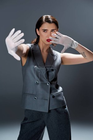 A stylish young woman is striking a pose in an elegant gray suit with a vest, complemented by white gloves, in a studio with a grey background.