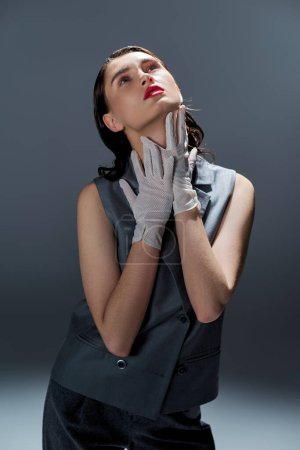 A stylish young woman in an elegant suit with a vest, hands on her face, showcasing raw emotions in a studio setting on a grey background.