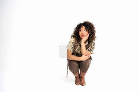 Photo for Alluring african american woman in stylish attire squatting on white backdrop and looking away - Royalty Free Image