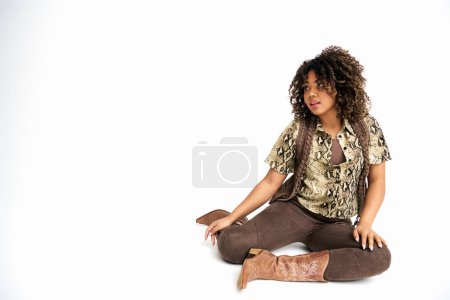 Foto de Mujer afroamericana con estilo en traje elegante sentado en el suelo sobre fondo blanco y mirando hacia otro lado - Imagen libre de derechos