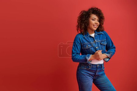 Photo for Joyous elegant african american woman in stylish denim outfit looking away on red backdrop - Royalty Free Image