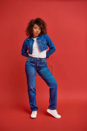 Photo for Attractive chic african american woman in denim attire posing on red backdrop and looking at camera - Royalty Free Image