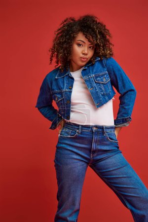 Photo for Elegant african american woman in denim attire posing on red backdrop and looking at camera - Royalty Free Image