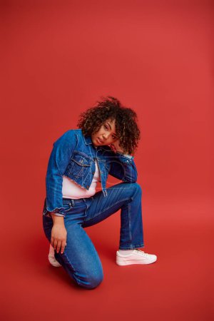 trendige afrikanisch-amerikanische Fashionista in Jeanskleidung posiert vor rotem Hintergrund und blickt in die Kamera