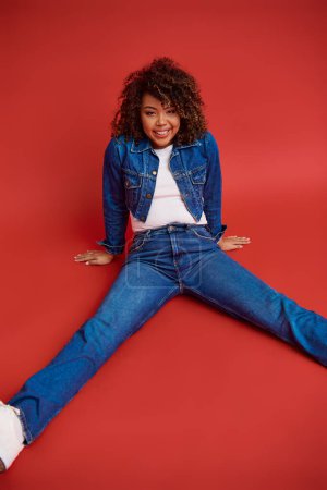 Photo for Joyous african american fashionista in denim attire posing on red backdrop and looking at camera - Royalty Free Image