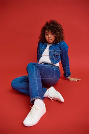 Photo for Young african american female model in denim attire looking at camera and posing on red background - Royalty Free Image