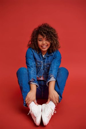 Photo for Joyful african american female model in denim attire looking at camera and posing on red background - Royalty Free Image