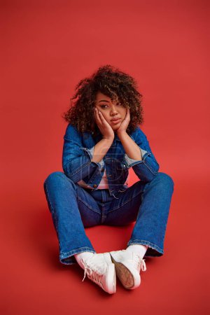 Photo for Beautiful young african american woman in stylish denim outfit looking away on red backdrop - Royalty Free Image