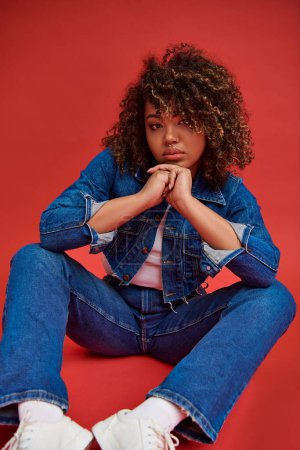 Photo for Fancy african american female model in denim attire looking at camera and posing on red background - Royalty Free Image