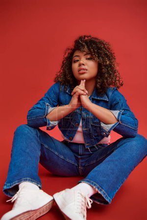 Photo for Alluring african american female model in denim attire looking at camera and posing on red backdrop - Royalty Free Image