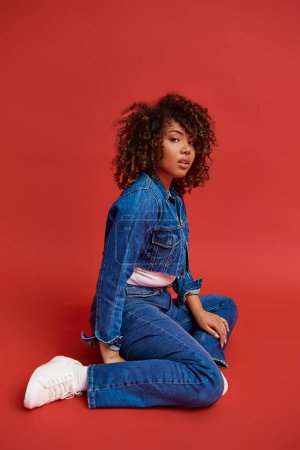 Photo for Refined african american woman in stylish denim outfit looking at camera on vibrant red backdrop - Royalty Free Image
