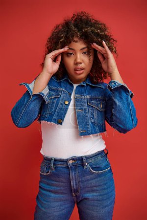 Photo for Graceful african american woman in stylish denim outfit looking at camera on vibrant red backdrop - Royalty Free Image
