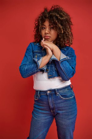 Photo for Well dressed african american woman in denim outfit looking at camera on vibrant red backdrop - Royalty Free Image