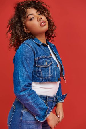 Photo for Appealing african american woman in stylish denim outfit looking at camera on vibrant red backdrop - Royalty Free Image