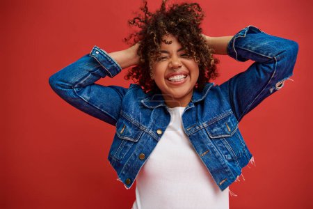 Photo for Cheerful african american woman in denim attire posing happily with closed eyes on red backdrop - Royalty Free Image