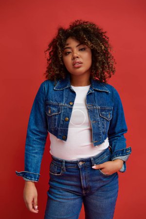 Photo for Alluring african american woman in stylish denim outfit looking at camera on vibrant red backdrop - Royalty Free Image