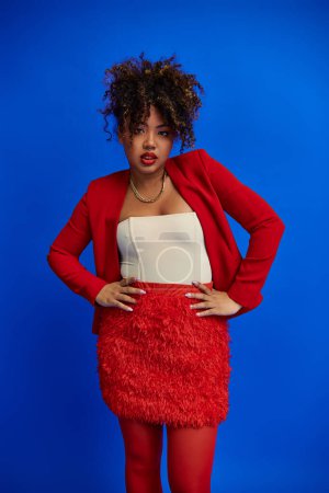 Photo for Trendy african american woman in red skirt and blazer posing on blue backdrop and looking at camera - Royalty Free Image