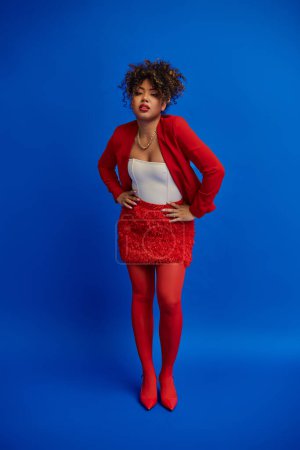 Photo for Stylish african american woman in red skirt and blazer posing on blue backdrop and looking at camera - Royalty Free Image