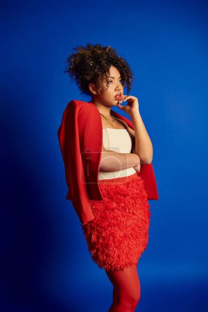 Foto de Exquisito modelo femenino afroamericano con estilo en traje vibrante mirando hacia otro lado en el fondo azul - Imagen libre de derechos