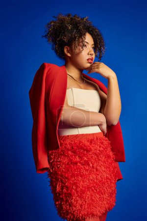 Téléchargez les photos : Attrayant modèle féminin afro-américain élégant en tenue vibrante regardant loin sur fond bleu - en image libre de droit