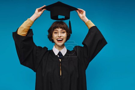 sonriente chica universitaria con vestido de graduación negro celebración de la tapa académica sobre fondo azul en el estudio
