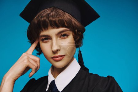 portrait of pensive college girl wearing black graduation gown and academic cap on blue backdrop