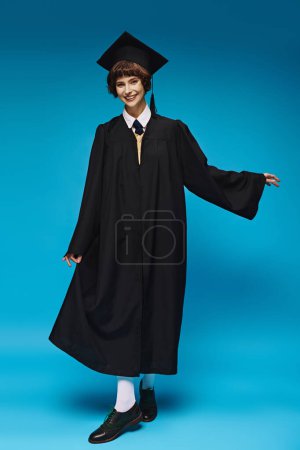 concepto de graduación, chica universitaria feliz en gorra académica y vestido de pie sobre fondo azul