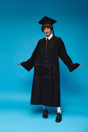 concepto de graduación, chica universitaria optimista en gorra académica y vestido de pie sobre fondo azul