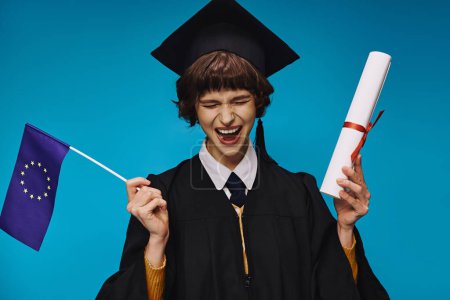 Photo for Excited graduated college girl in gown and academic cap with diploma and EU flag on blue backdrop - Royalty Free Image